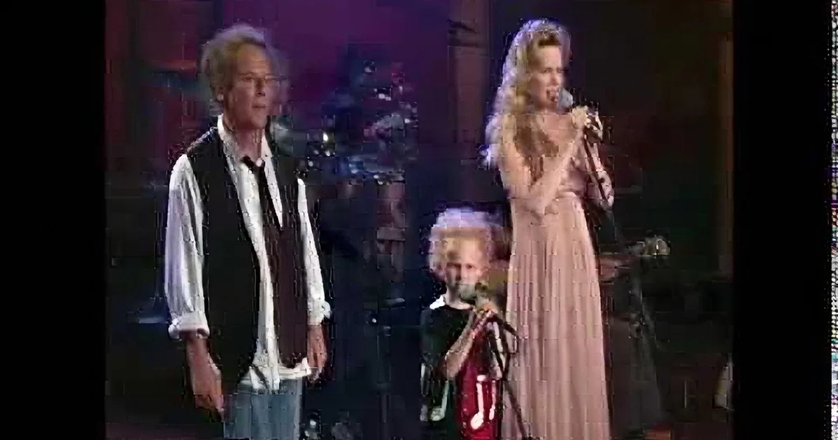A man and a young boy sing on stage, both holding microphones, with a keyboardist in the background.