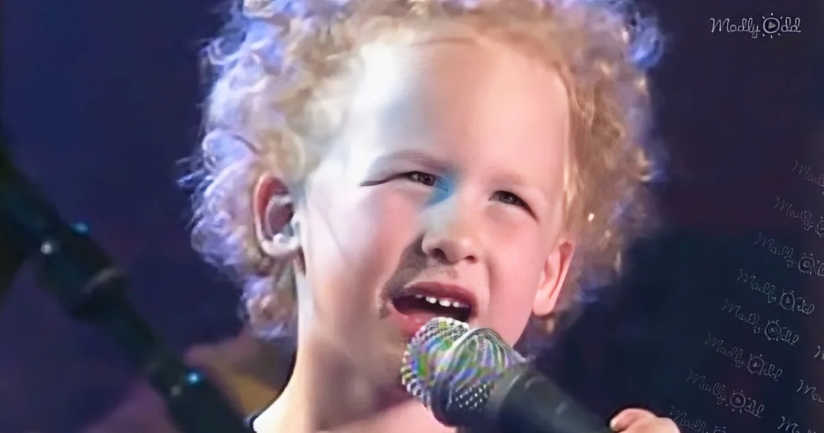 A young child with curly blonde hair sings into a microphone on stage.