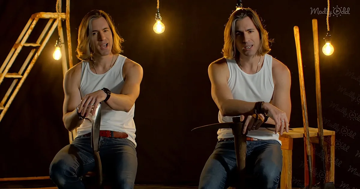 A man in a white tank top and jeans sits on a stool, holding a knife, with a dark background and hanging light bulbs.