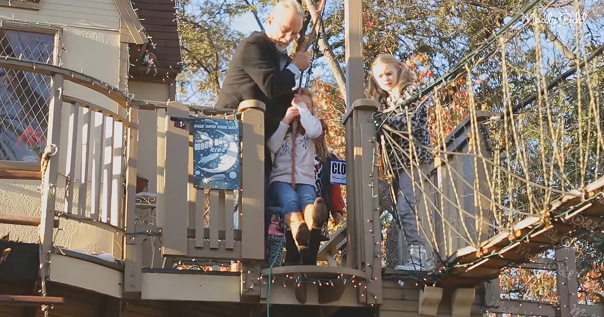 A man helps a child on a swing at a treehouse playground with festive lights.