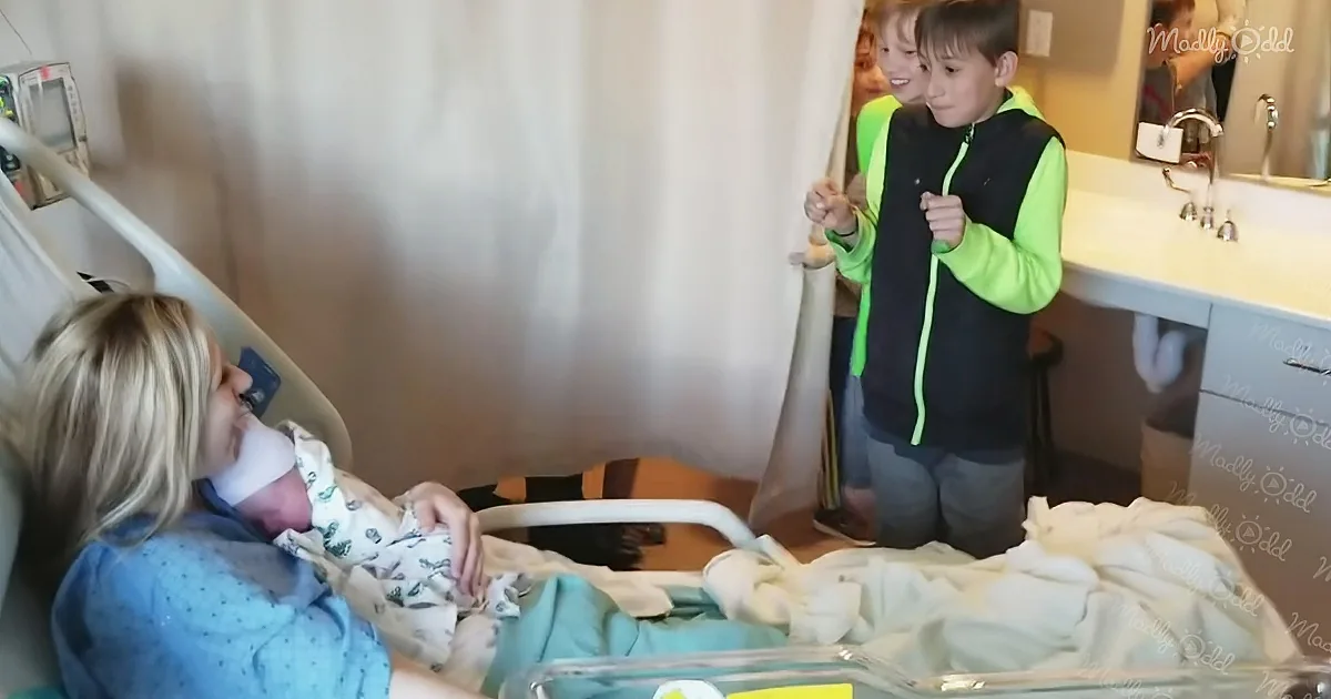 A woman holds a newborn baby in a hospital bed, with two children peeking from behind a curtain nearby.