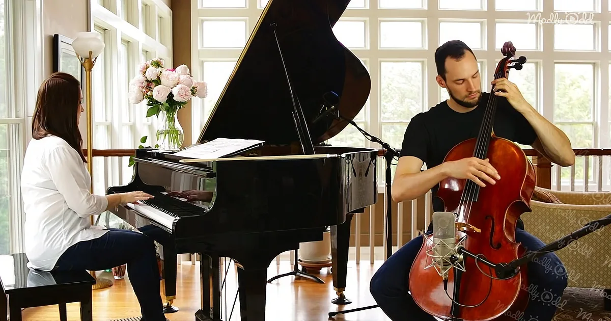 A woman plays piano while a man plays cello in a bright room with large windows.
