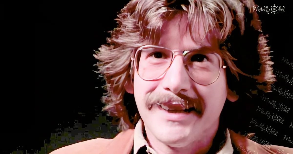 A person with curly hair, glasses, and a mustache is shown in a close-up against a dark background.