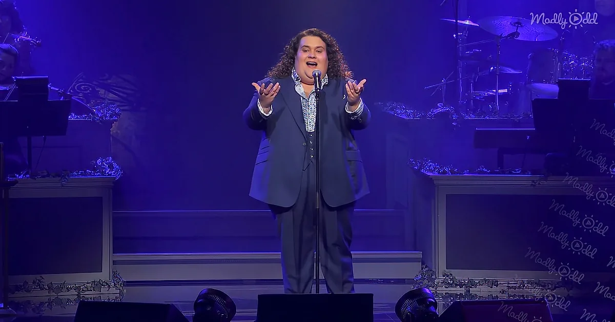 A man in a suit sings passionately on stage with a microphone, surrounded by musical instruments and purple lighting.