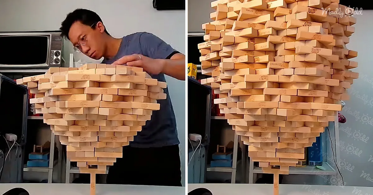 A man is building a tall, intricate tower of wooden blocks, resembling a cone shape, in a room with shelves and a microwave.