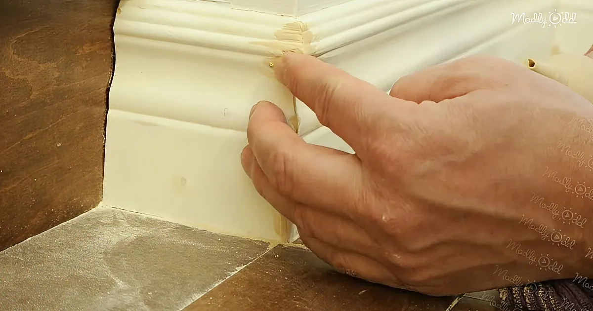 A hand is carefully applying a small piece of material to a corner of a white baseboard.