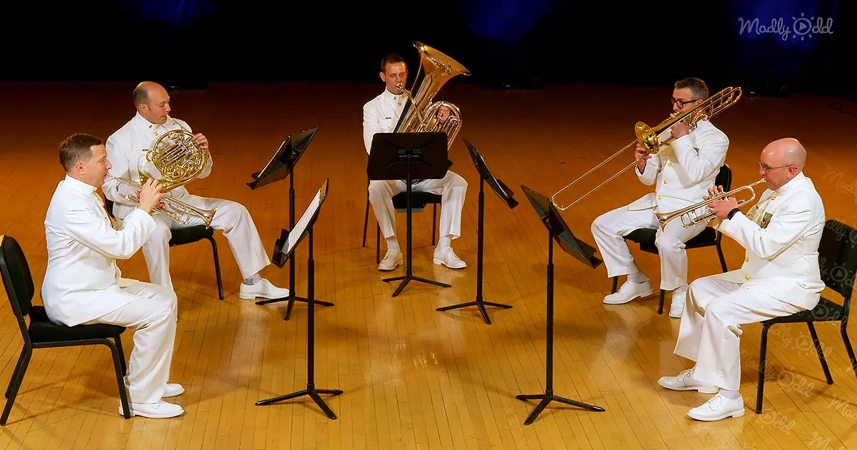 Five men in white uniforms play brass instruments on stage with music stands.