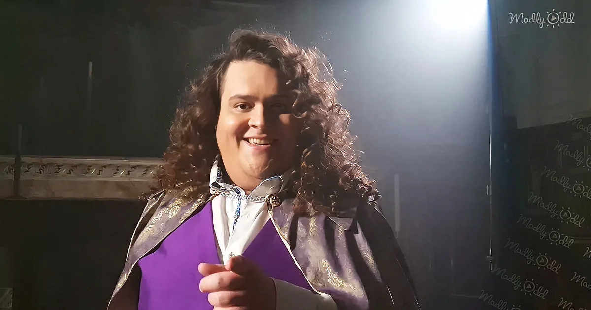 A person with curly hair, wearing a purple vest and white shirt, smiles in a dimly lit room with a spotlight.