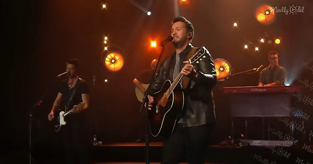 A man in a black leather jacket plays an acoustic guitar on stage, illuminated by warm lights.