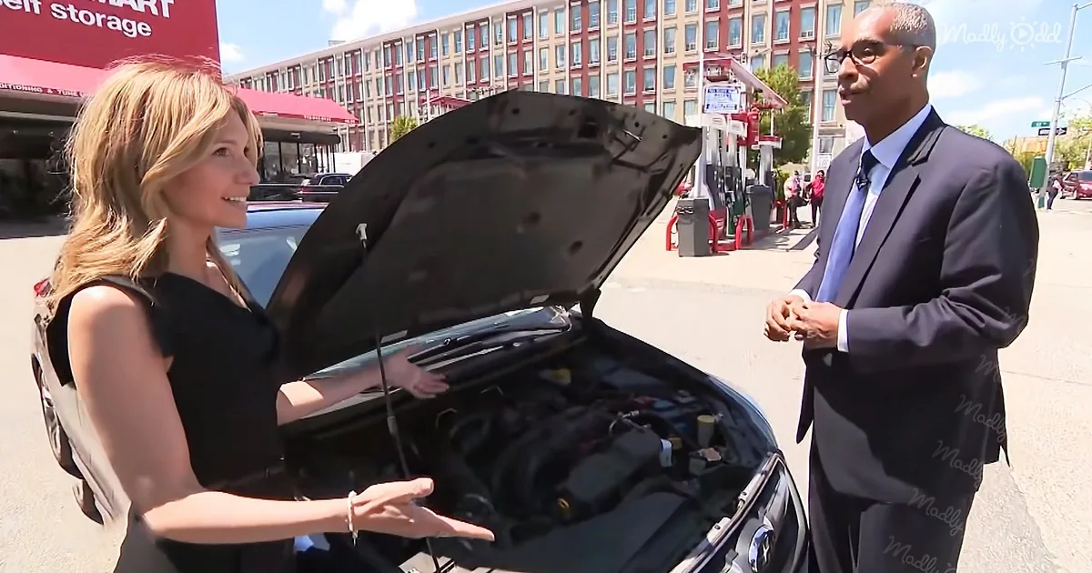 A woman in a black dress opens a car's hood, while a man in a suit stands nearby, both in an urban setting.