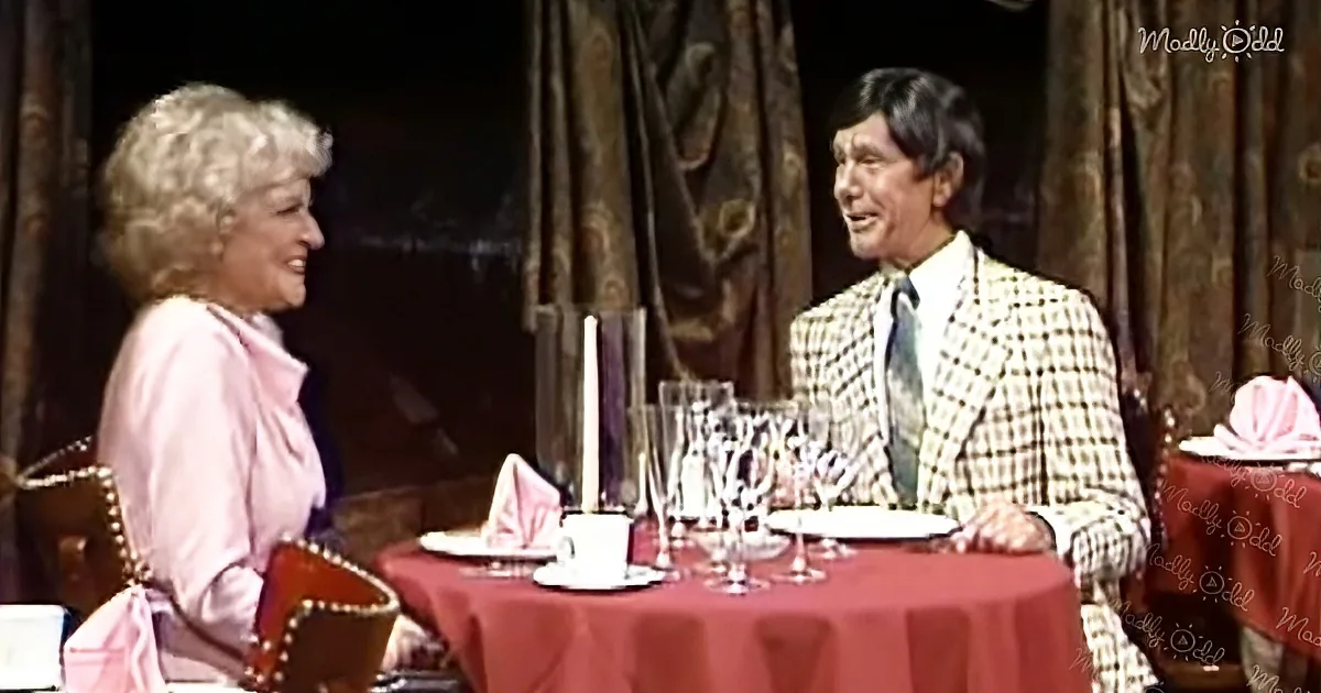 A woman in pink and a man in a checkered suit sit at a table with red tablecloth, smiling and talking.