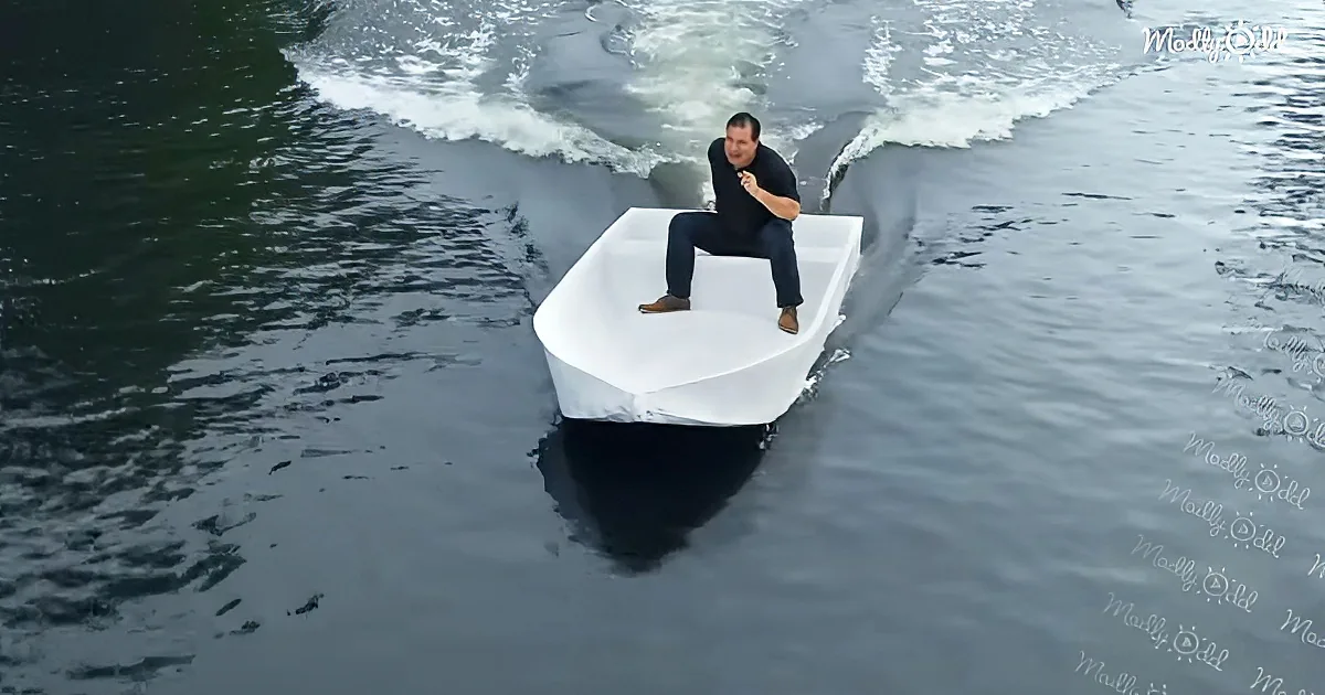 A man sits on a white, paper-like boat in the water, creating a wake behind him.