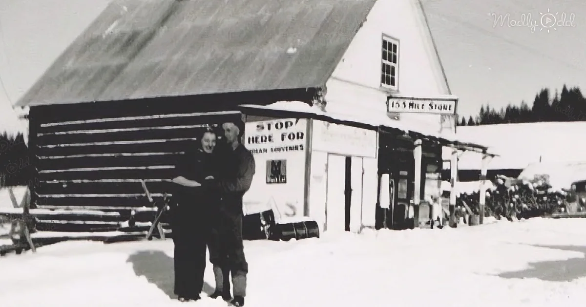 Two people stand in front of a snow-covered building labeled "153 Mile Store," with a sign advertising souvenirs.