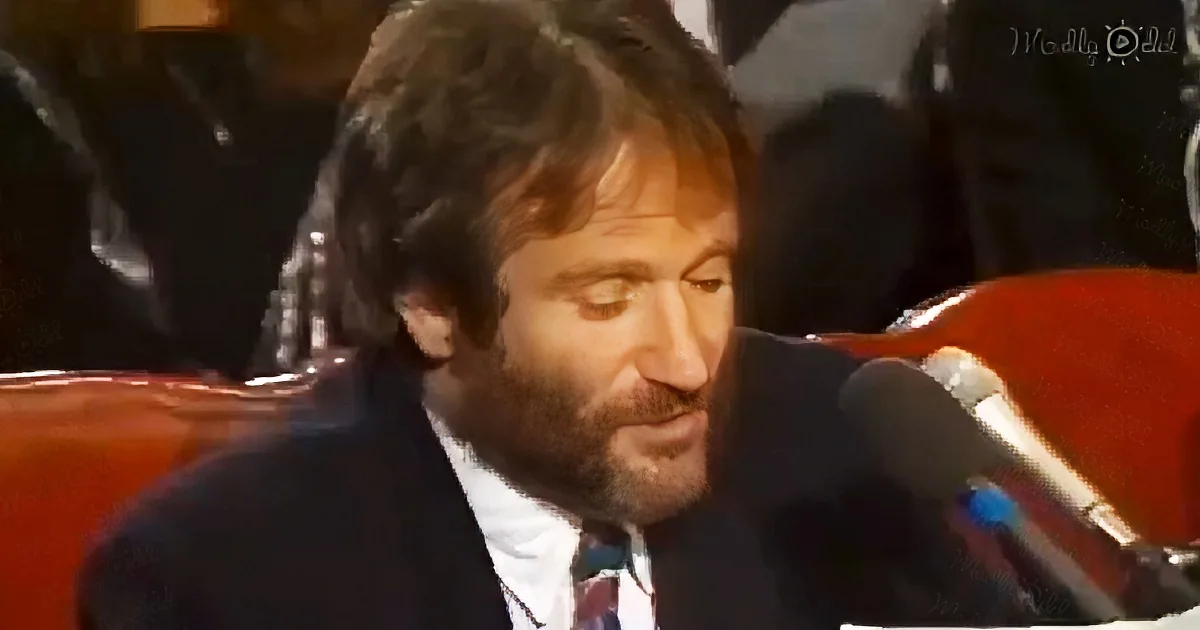 A man with a beard and dark hair is speaking into a microphone, wearing a suit and tie.