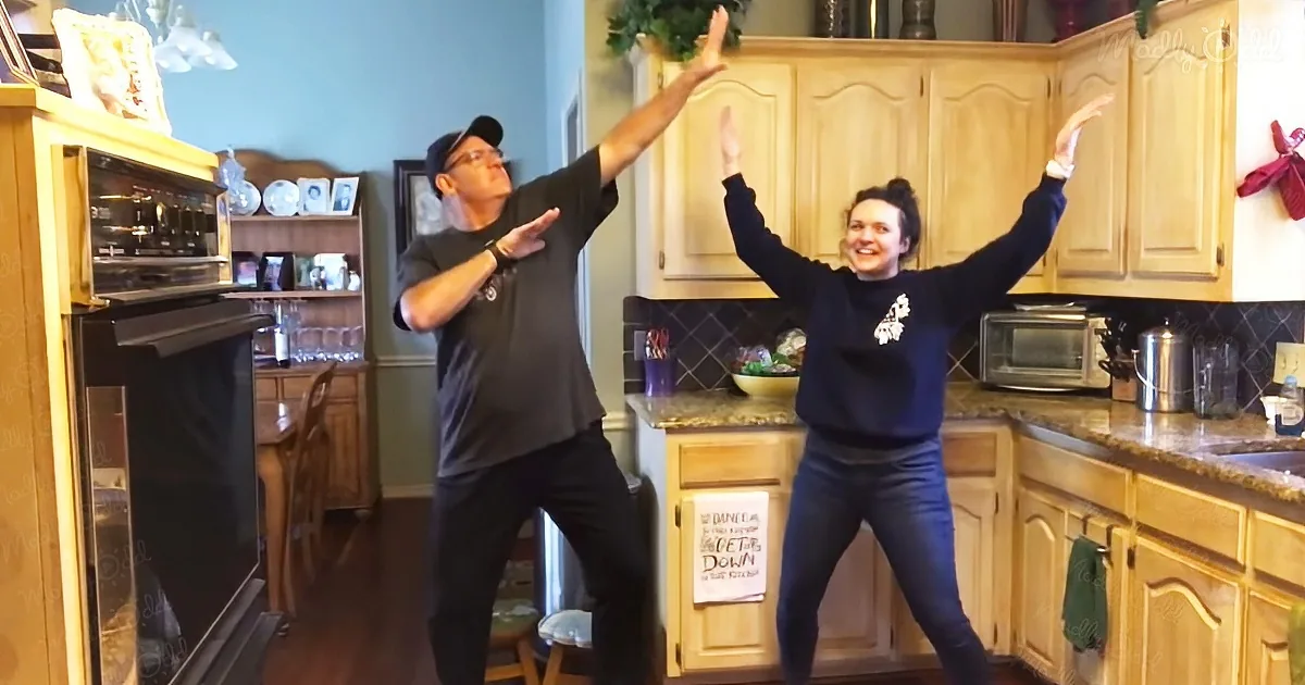 A man and woman dance joyfully in a kitchen, arms raised, smiling.