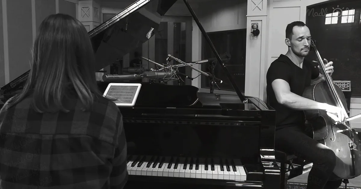 A man plays cello while a woman reads sheet music on a piano in a recording studio.