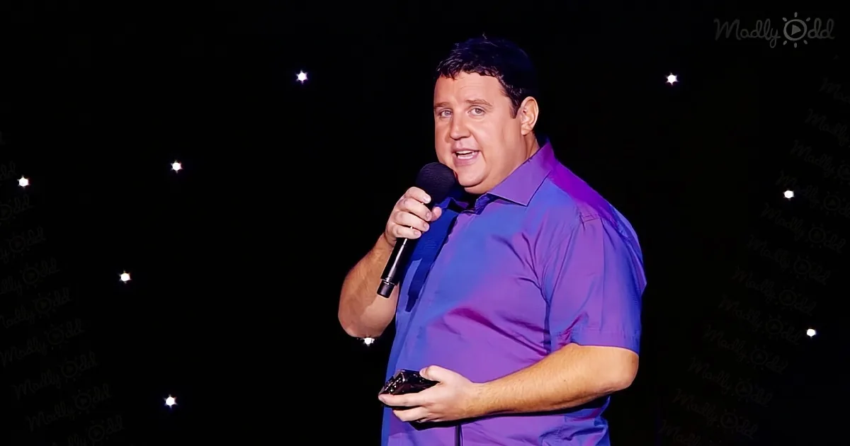 A man in a purple shirt holds a microphone, smiling, with a dark background and twinkling lights.