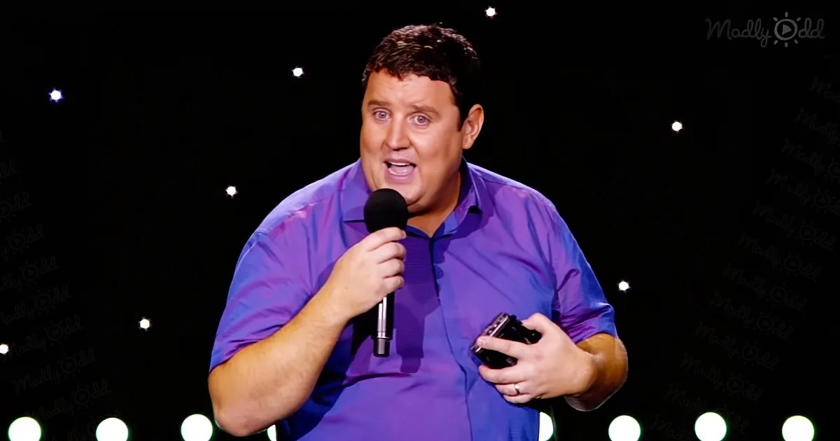 A man in a purple shirt holds a microphone, speaking on stage with a dark background and twinkling lights.