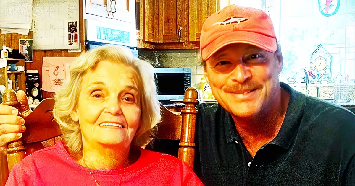 An older woman in a red shirt and a man in a black shirt and red cap are smiling in a kitchen.