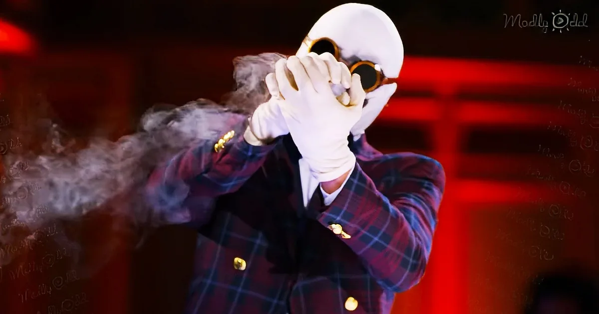 A person in a plaid suit and white gloves holds a mask, with smoke billowing from their hand.