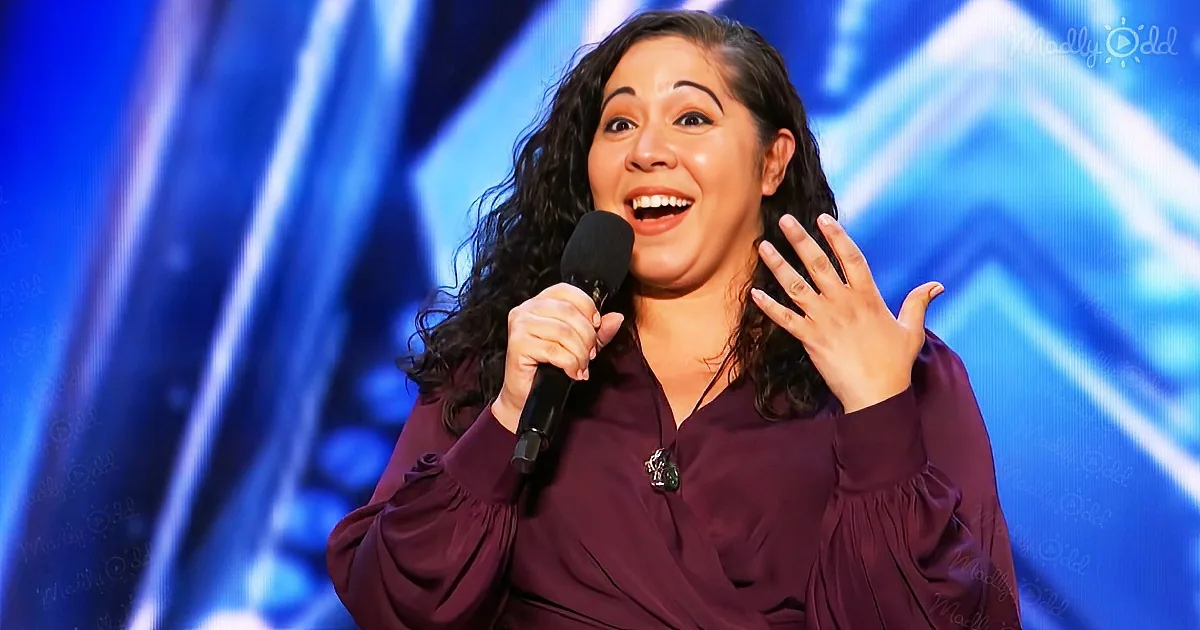 A woman in a maroon blouse holds a microphone, smiling with her hand raised, on a stage with blue lighting.
