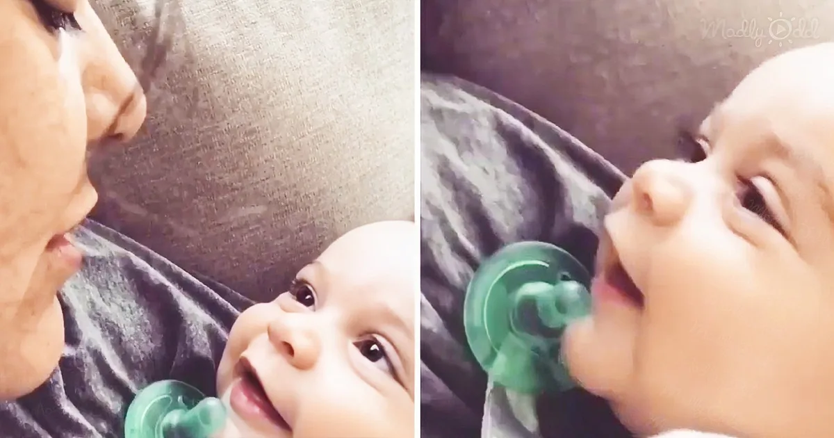 A baby with a green pacifier is lying on a gray blanket, looking up at a person who is partially visible.