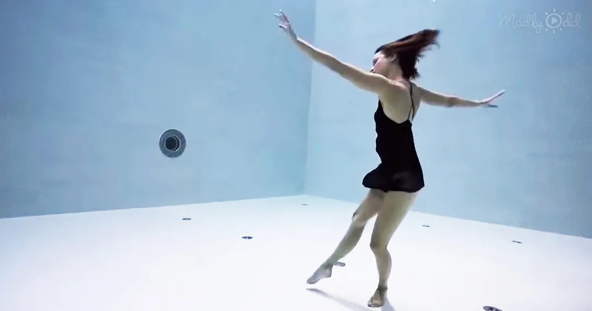 A woman in a black dress dances gracefully underwater in a pool.