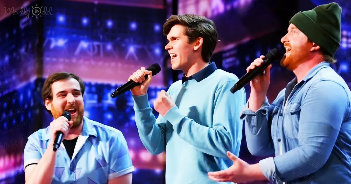 Three men in blue shirts sing into microphones on a stage with a colorful background.