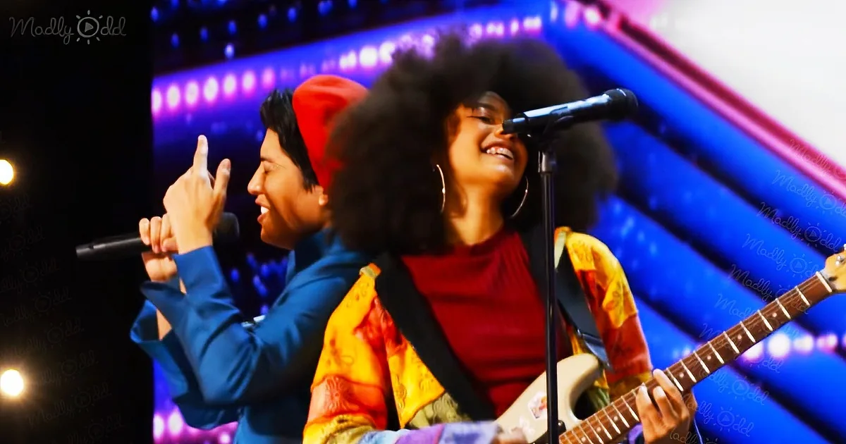 A woman with an afro plays guitar while another person sings into a microphone on stage.