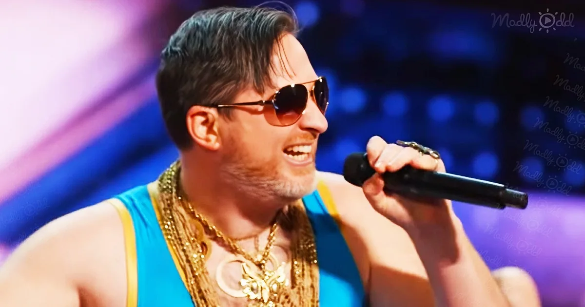 A man in a blue tank top and sunglasses is singing into a microphone on stage.