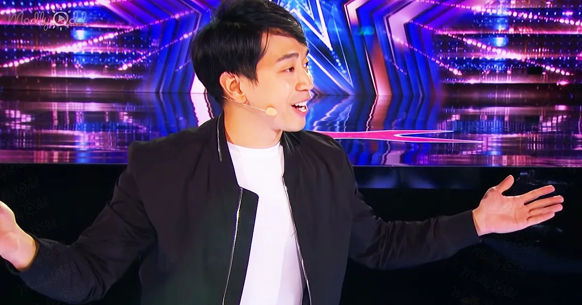 A man in a black jacket and white shirt gestures with his hands on a stage with colorful lights.