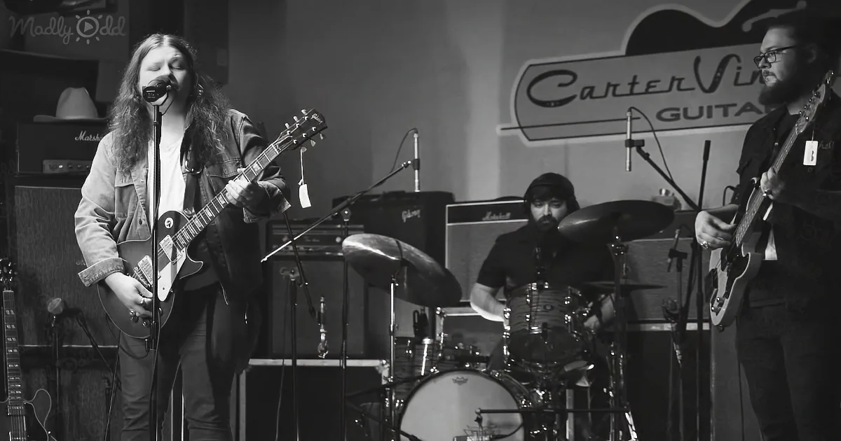A musician with long hair plays guitar and sings into a microphone on stage.