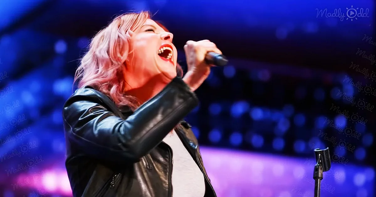 A person in a leather jacket sings passionately into a microphone on stage with vibrant blue and purple lighting.
