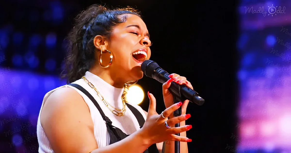 A woman in a white and black outfit sings passionately into a microphone, adorned with gold jewelry and red nails.