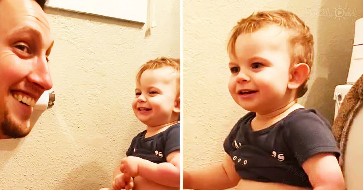 A smiling baby in a dark shirt holds a finger, with a man in the background.