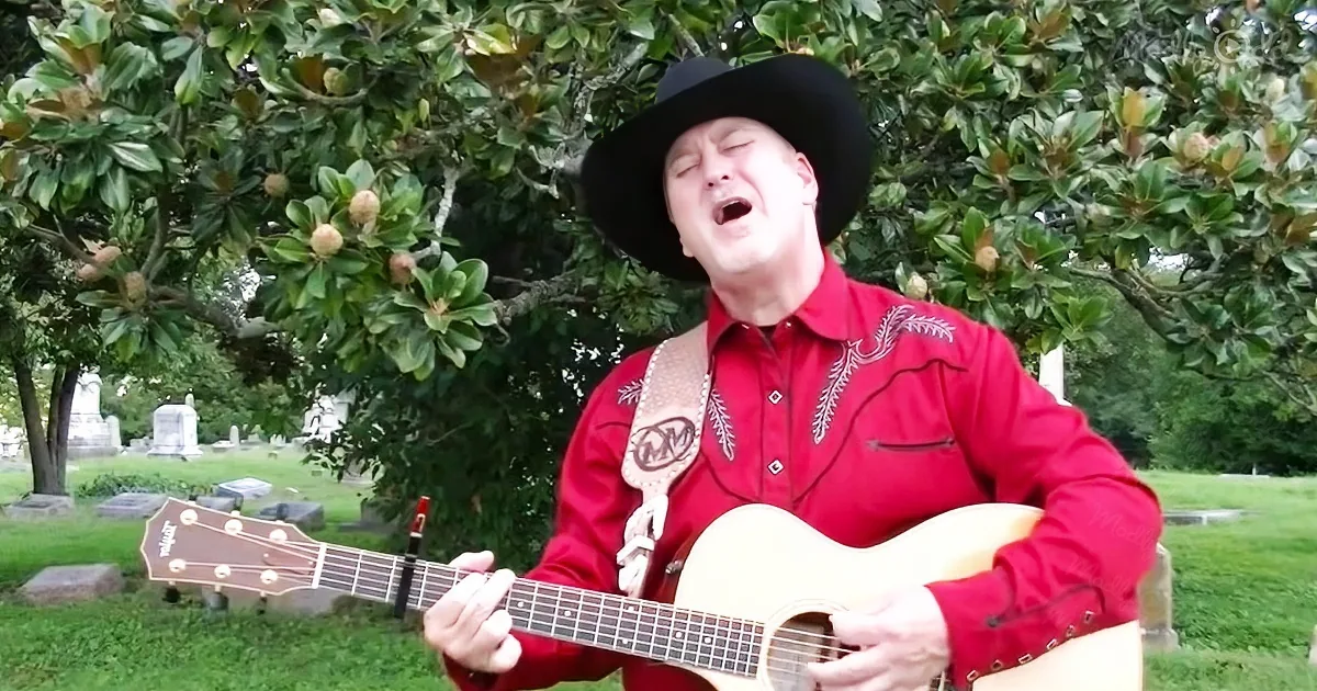 A man in a red shirt and cowboy hat sings while playing an acoustic guitar in a cemetery.