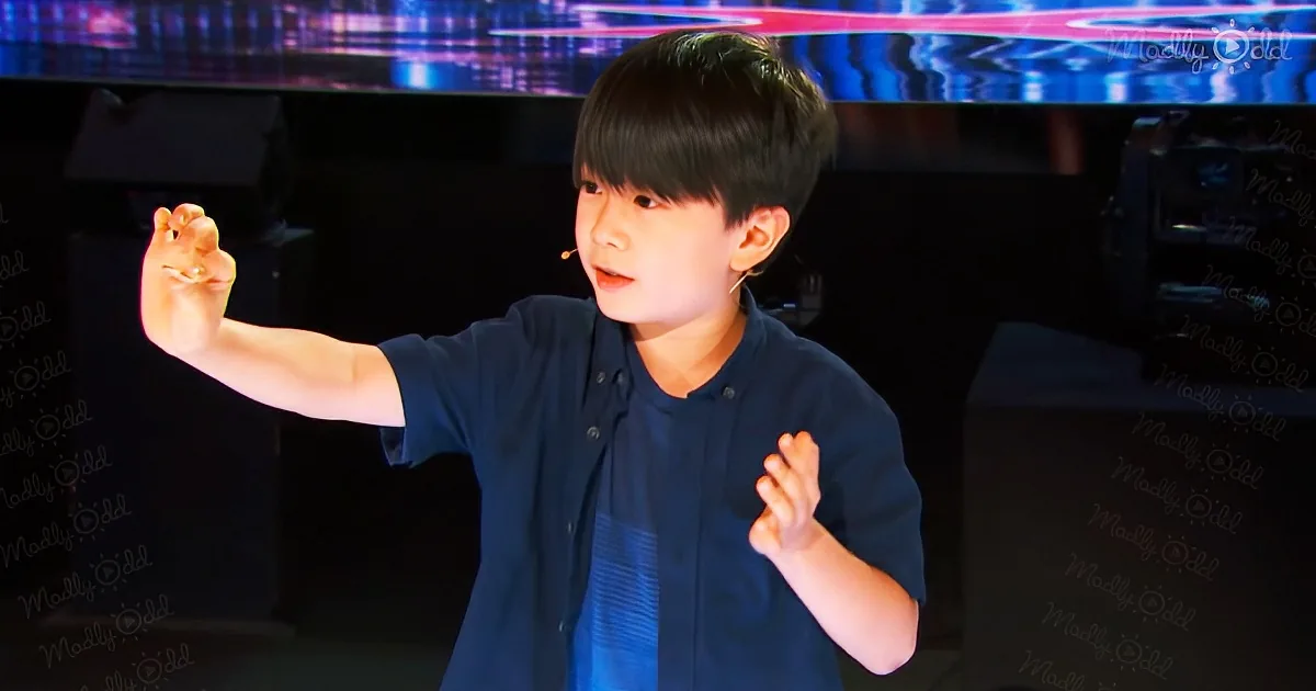 A young boy in a dark shirt gestures with his hands on stage, wearing a microphone.