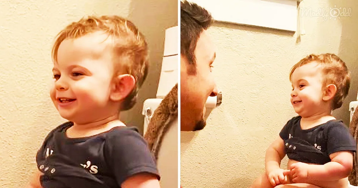 A smiling toddler in a dark shirt sits near a wall, interacting with an adult.