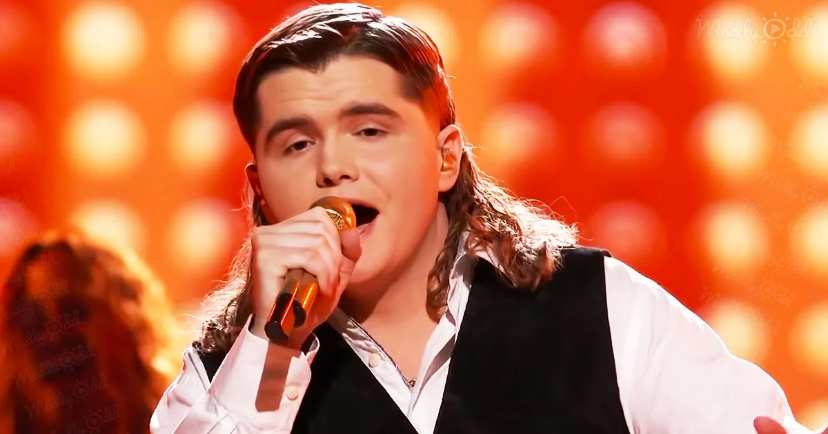 A person with long hair, wearing a white shirt and black vest, sings into a microphone on stage with a red background.