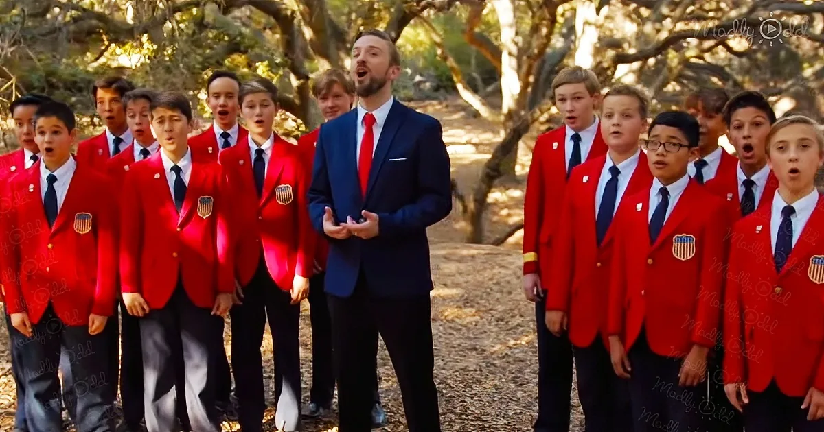 A man in a blue suit leads a group of boys in red jackets singing outdoors.
