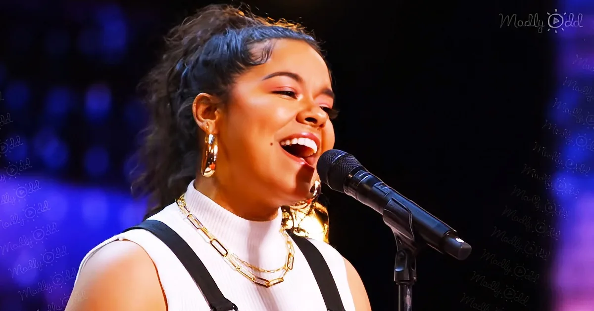 A woman sings into a microphone, wearing a white top and gold necklace, with a dark background.