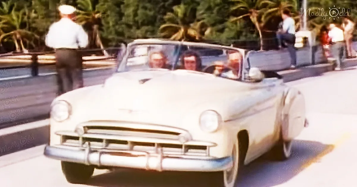 A vintage white convertible car is parked on a street with palm trees and people in the background.