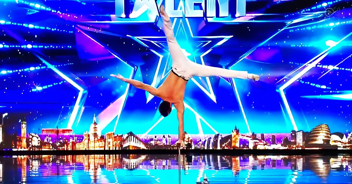 A man performs a handstand on a pole in front of a cityscape backdrop on a stage.