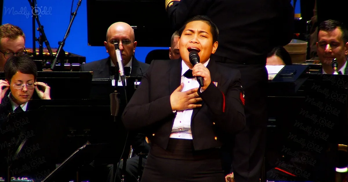 A person in a formal outfit sings into a microphone, surrounded by musicians and instruments.