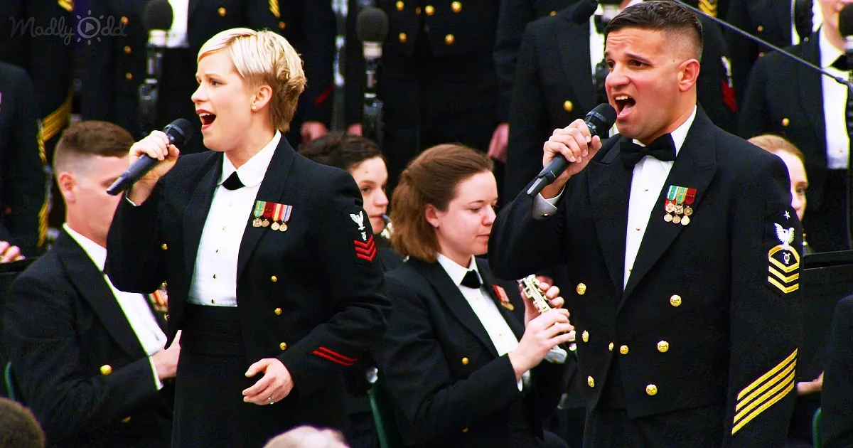 A man and woman in military uniforms sing into microphones, surrounded by other uniformed individuals.