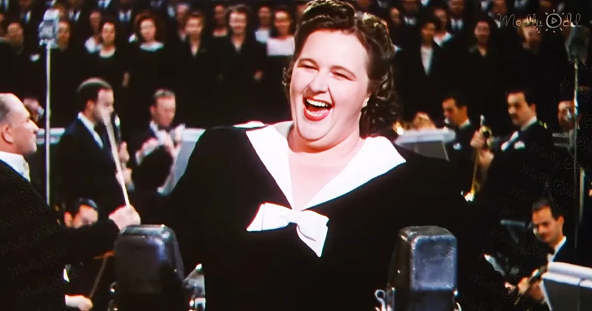 A woman in a sailor-style outfit sings into a microphone, surrounded by a band and an audience.