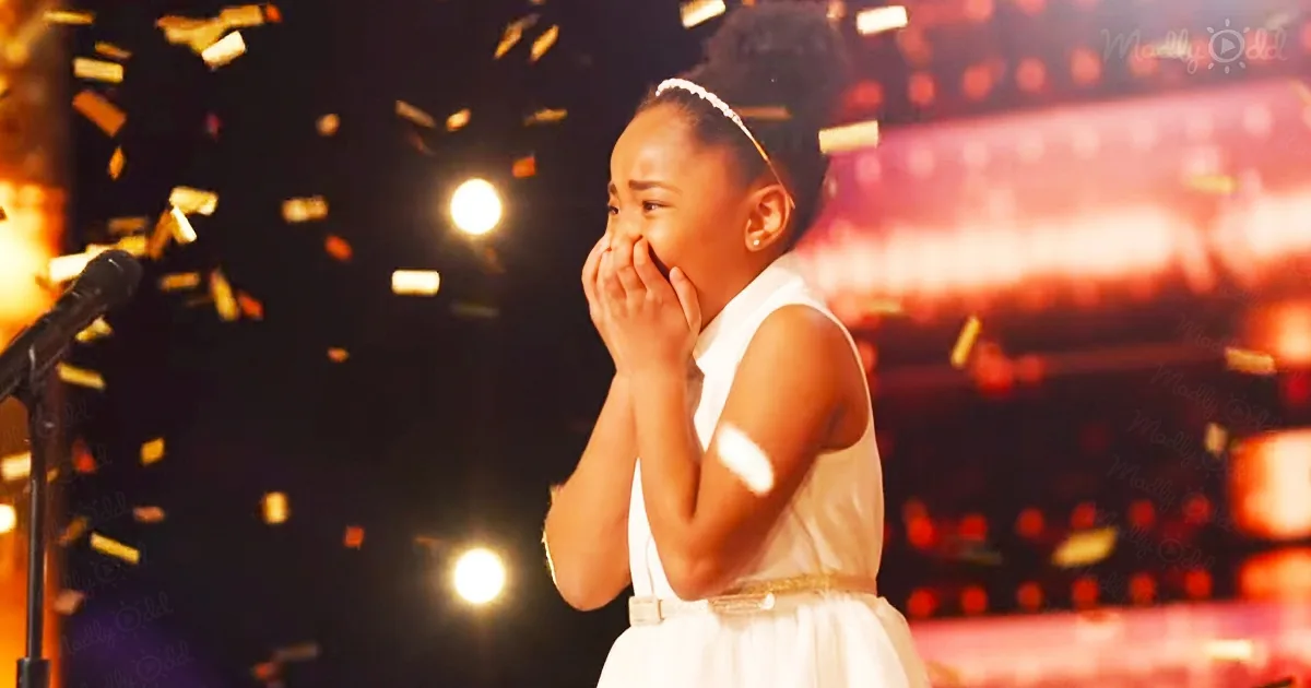 A young girl in a white dress covers her mouth, surrounded by confetti, on a stage with bright lights.