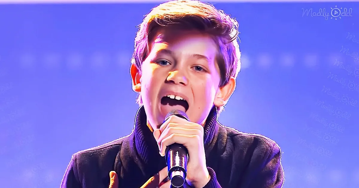 A young person sings passionately into a microphone, wearing a dark jacket, against a blue background.
