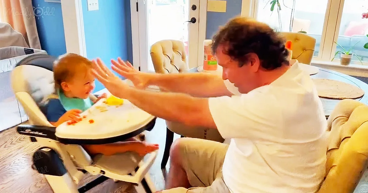 A man in a white shirt is playfully interacting with a baby in a high chair, both appearing amused.