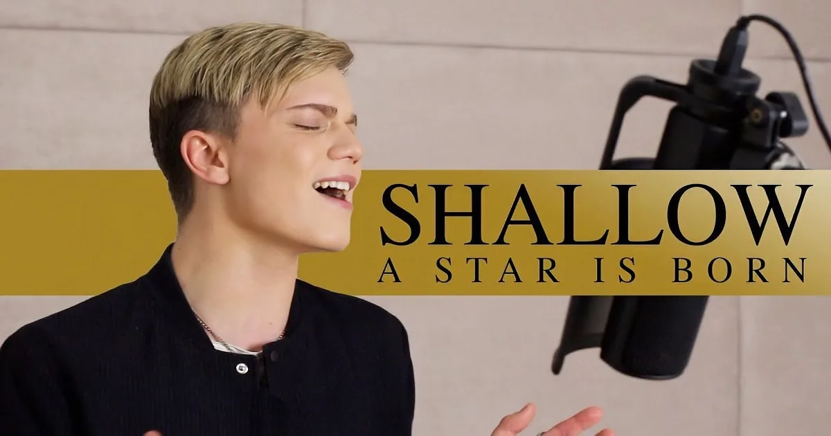 A person with short, light-colored hair wearing a black shirt is singing into a microphone.